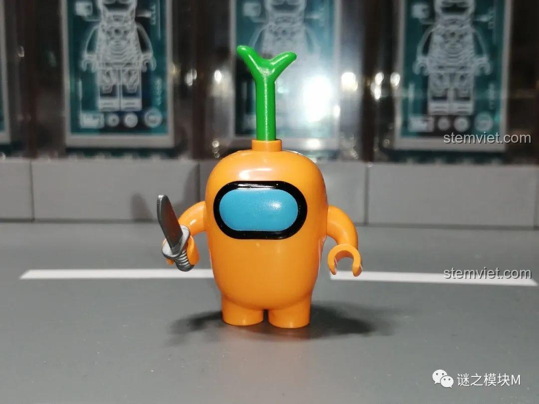 Phi hành gia Cam trong set đồ chơi Among Us PRCK 69702, cầm dao, với chi tiết lá cây trên đầu, rất dễ thương.