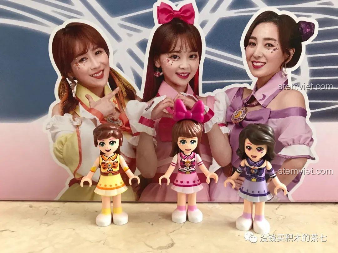 Ba minifigures Hạ Thiên, Tiểu Linh, Duyệt Nhi đứng trước hình ảnh trên hộp.