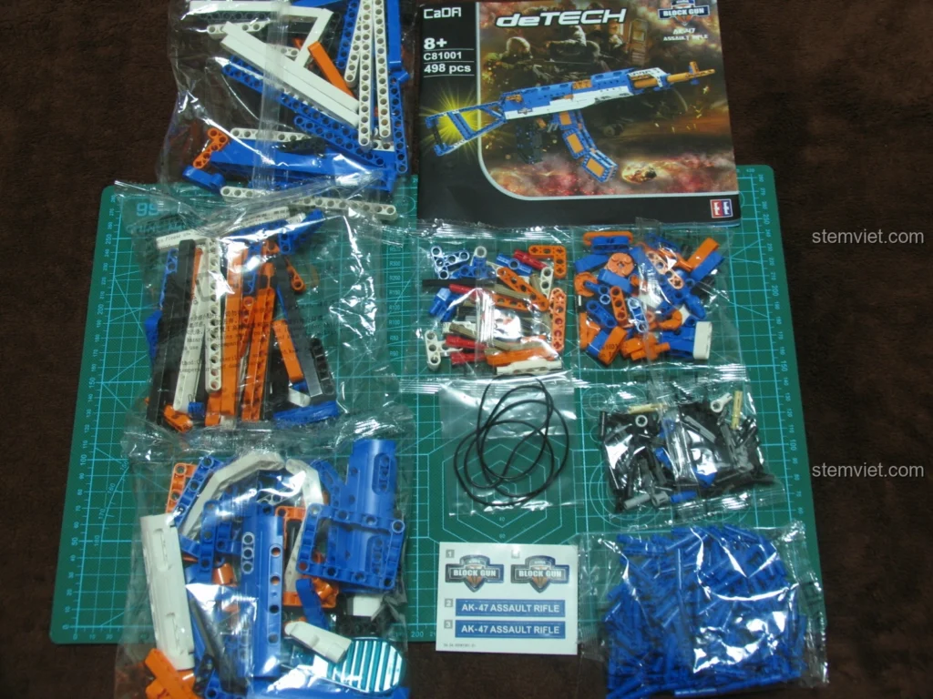 Bộ lắp ráp Súng trường tấn công AK-47 CaDA C81001 unbox, hiển thị các túi mảnh, dây chun, sticker và sách hướng dẫn trên thảm cắt.