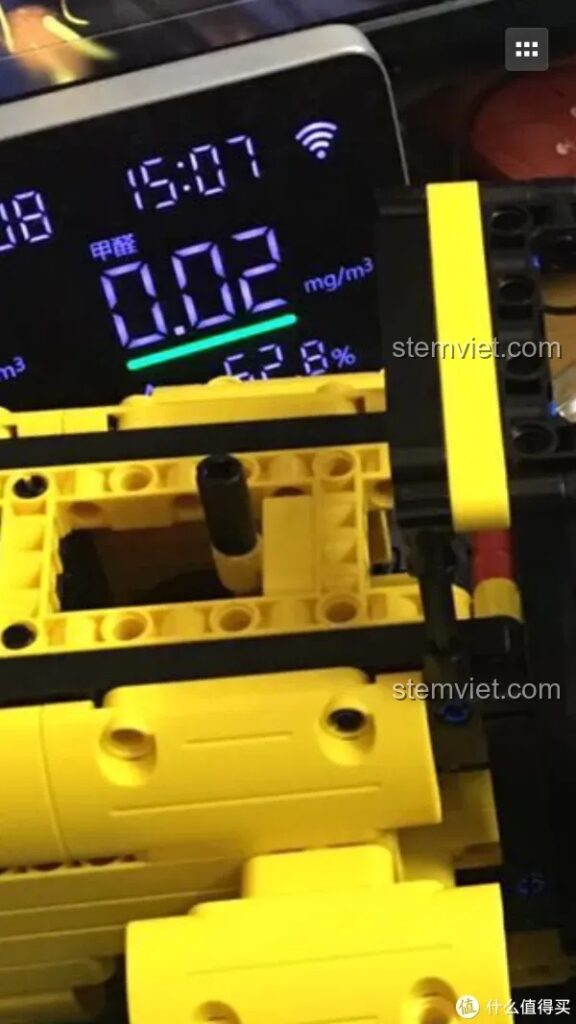 Cận cảnh màn hình hiển thị của máy đo chất lượng không khí với chỉ số 0.02 mg/m³ và 62.8%, đặt cạnh các mảnh ghép màu vàng của bộ lắp ráp xe xúc lật.