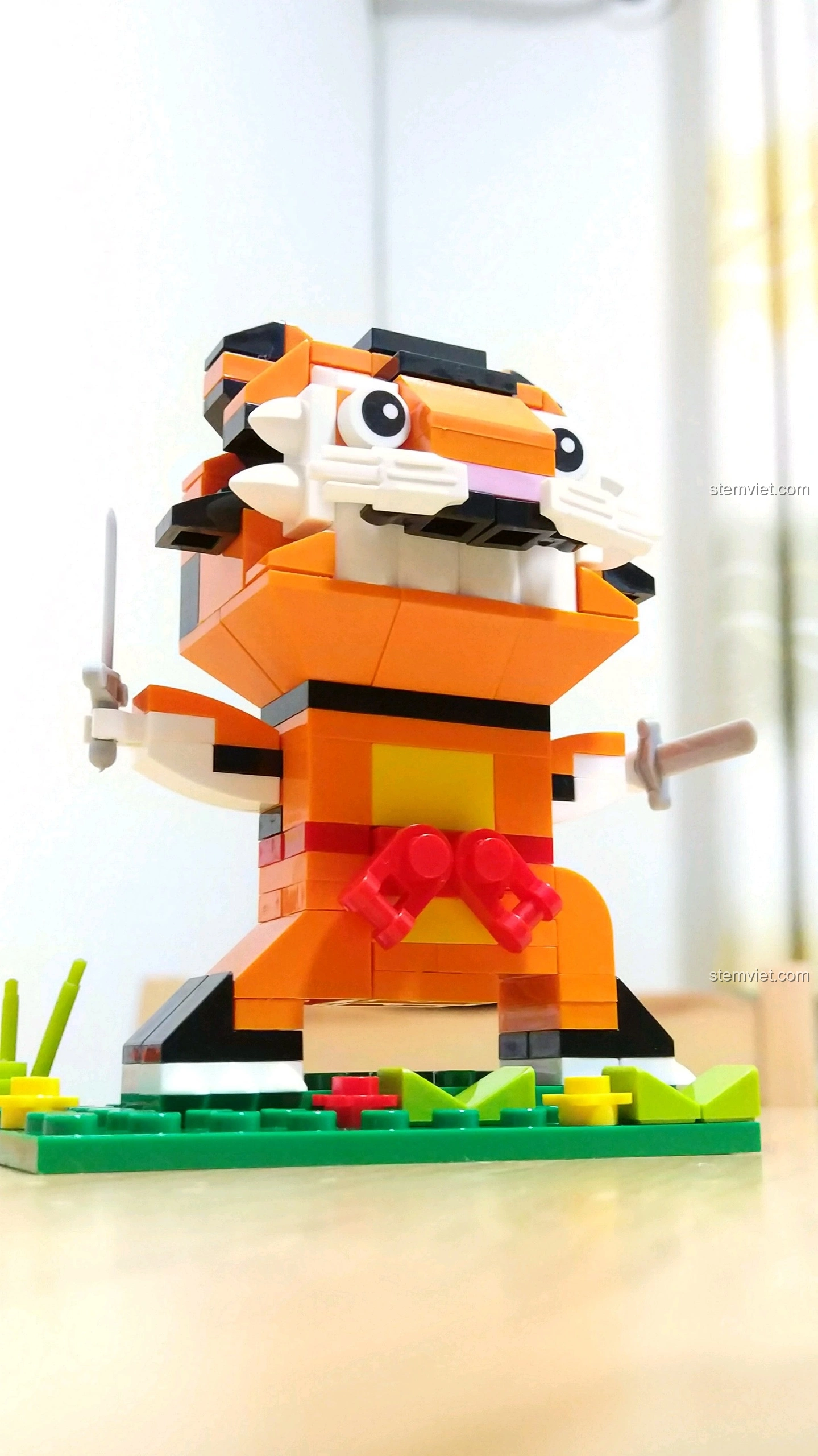 Cận cảnh mô hình Hổ Dần, tập trung vào khuôn mặt với răng nanh và hai thanh kiếm nhỏ, chi tiết trên nền cỏ xanh.