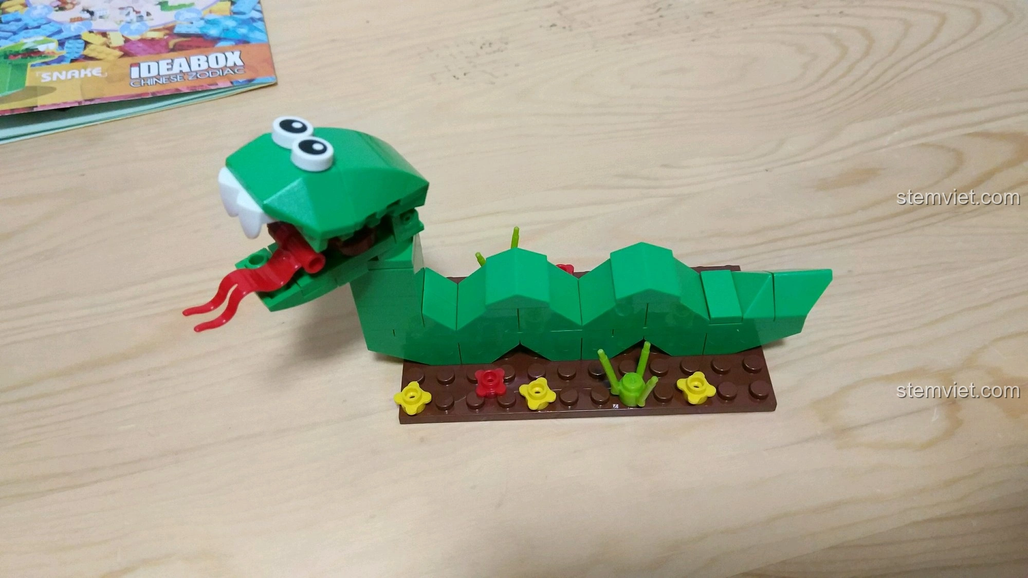 Toàn cảnh mô hình Rắn Tỵ màu xanh lá cây, thân uốn lượn trên nền đất nâu với các chi tiết hoa và cỏ nhỏ.