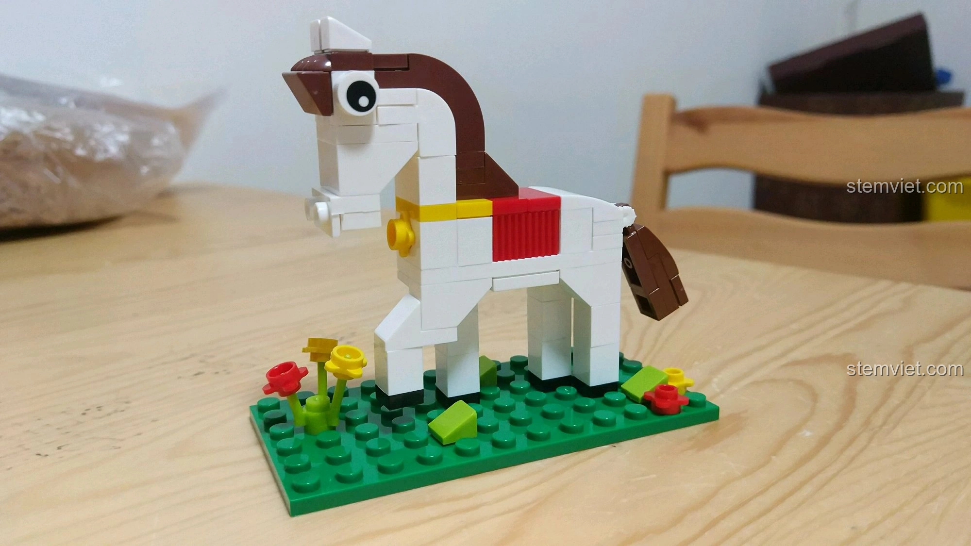 Mô hình Ngựa Ngọ màu trắng với yên đỏ và bờm nâu, đứng trên nền cỏ xanh có hoa, từ bộ lắp ráp 12 Con Giáp XINGBAO XB-18001.