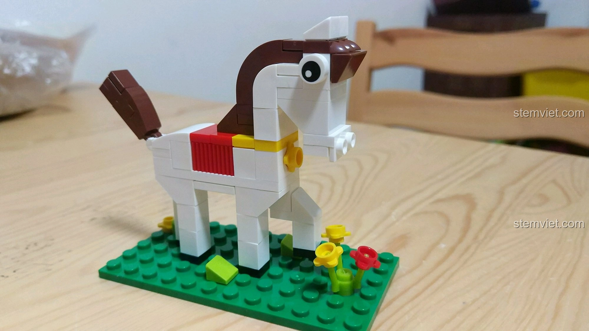 Góc nghiêng của mô hình Ngựa Ngọ, cho thấy chi tiết yên đỏ và bờm nâu, đứng trên nền cỏ xanh.