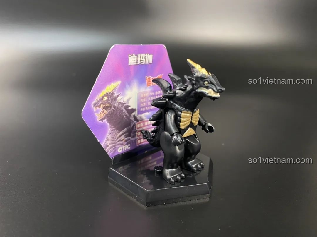 Figure quái vật Demaaga được đặt trên đế trưng bày hình lục giác với thẻ bài phía sau.