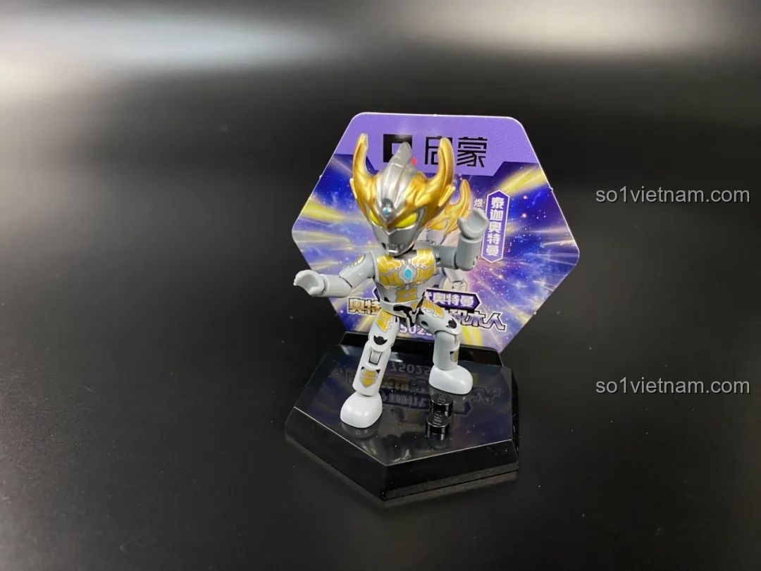 Figure Ultraman Taiga Tri-Strium được đặt trên đế trưng bày hình lục giác với thẻ bài phía sau.