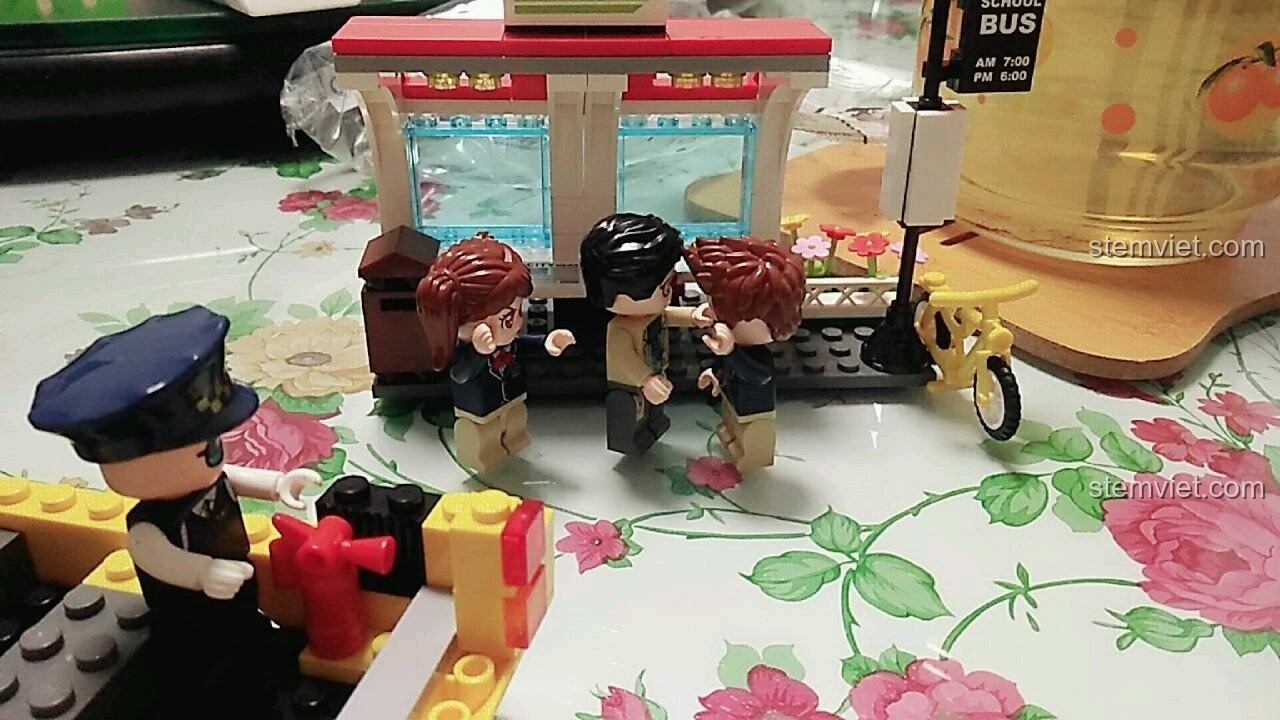 Cô giáo đang can thiệp khi một học sinh nam nghịch ngợm với xe đạp, gần trạm chờ xe buýt trong bộ QMAN 1136.