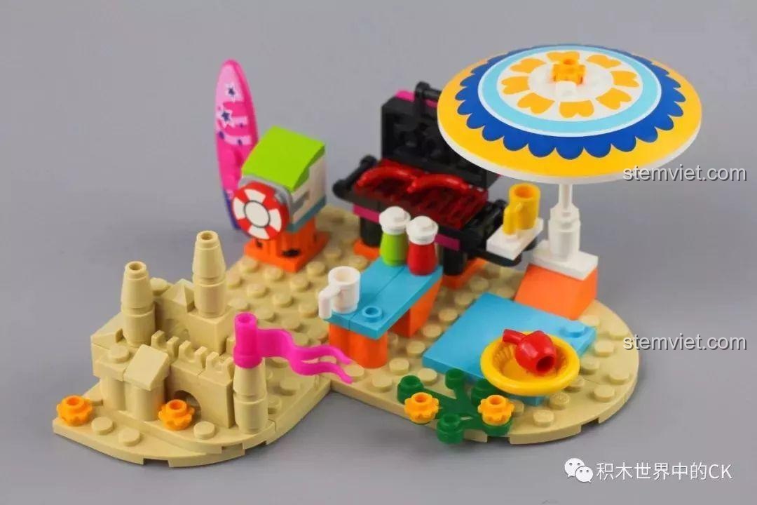 Toàn cảnh khu vực dã ngoại trên bãi biển của bộ QMAN 2018, bao gồm lâu đài cát, bếp nướng, bàn ăn và ván lướt sóng.