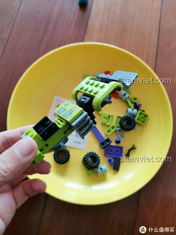 Bàn tay đang lắp ráp phần dưới của xe xúc, một trong những phương tiện của bộ Robot Biến Hình Đại Lực Thần SEMBO 103081, với các mảnh ghép màu xanh lá và tím.