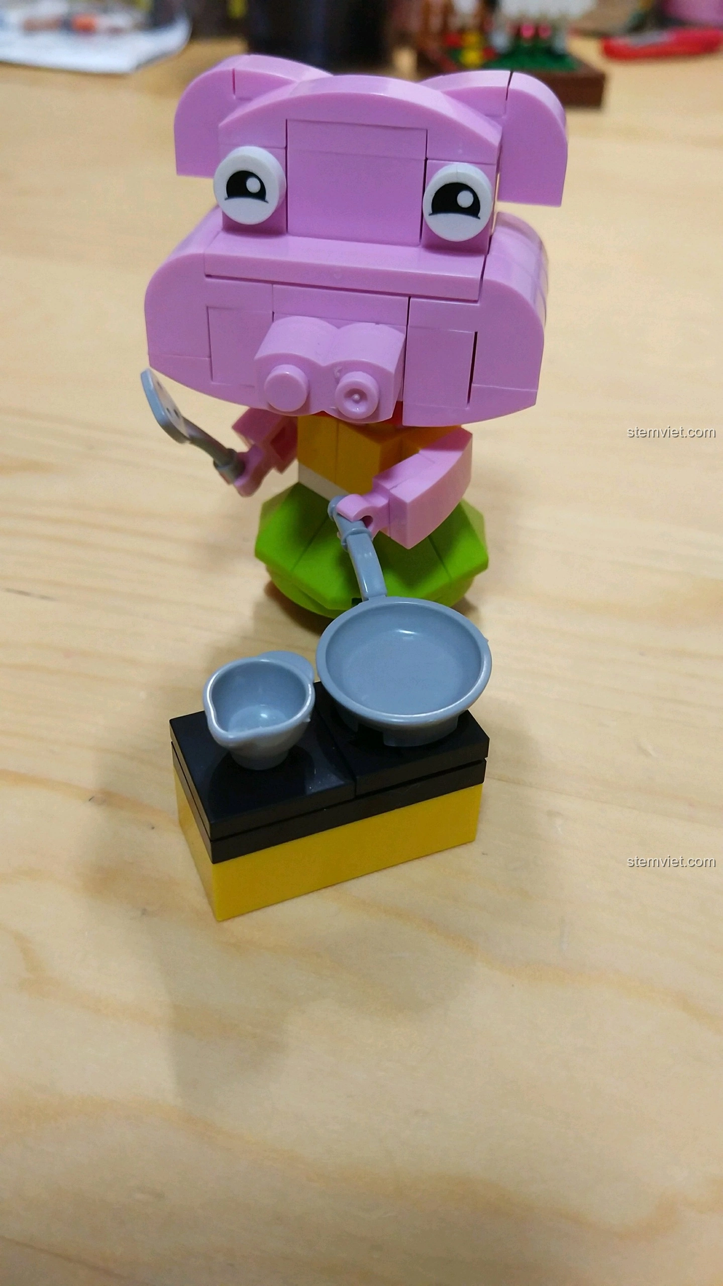 Mô hình Heo Hợi màu hồng với váy xanh, cầm xẻng và chảo, đứng cạnh bếp mini, từ bộ lắp ráp 12 Con Giáp XINGBAO XB-18001.