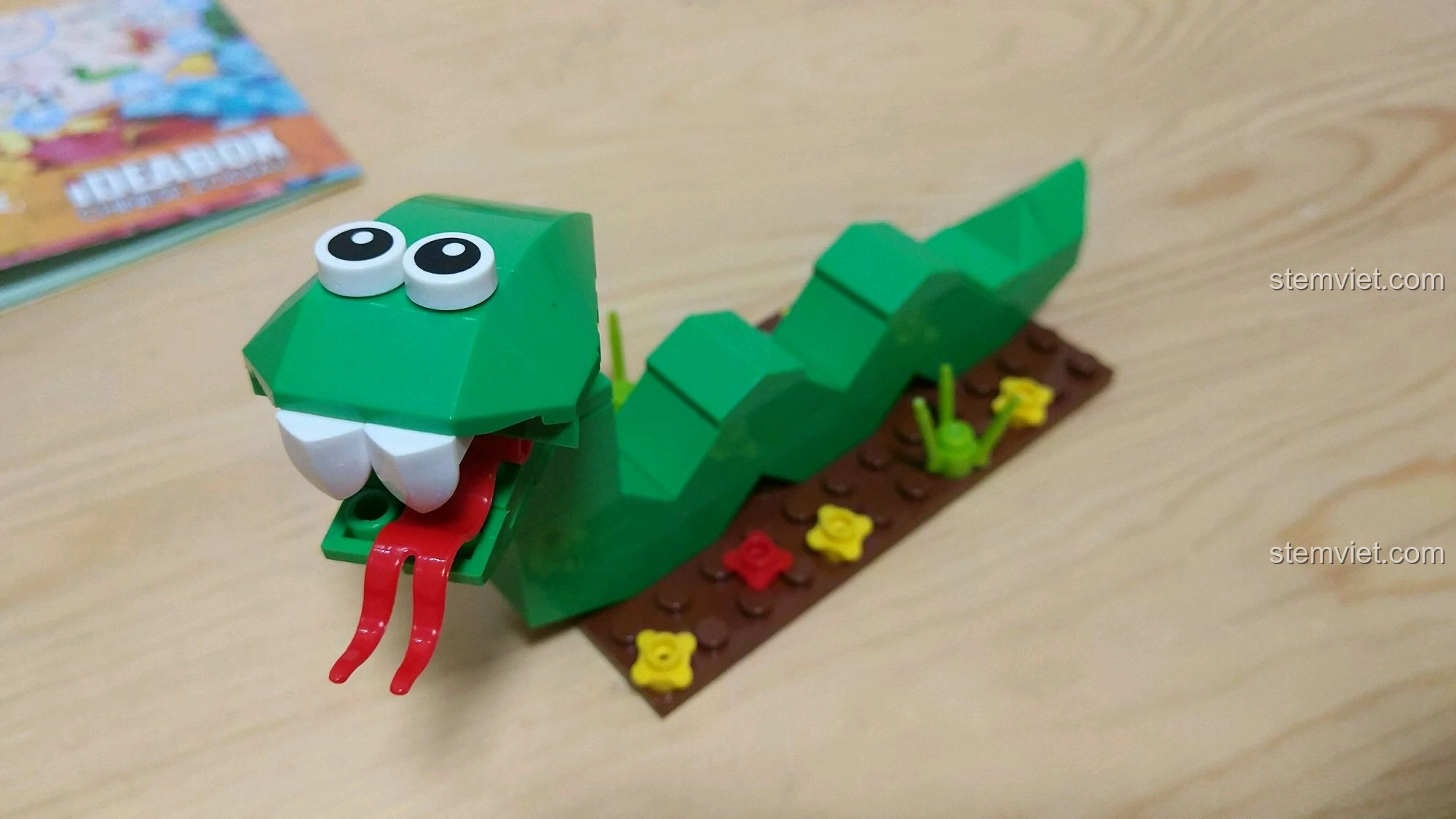 Mô hình Rắn Tỵ màu xanh lá cây với lưỡi đỏ chẻ đôi, đặt trên nền đất nâu có hoa nhỏ, từ bộ 12 Con Giáp XINGBAO XB-18001.