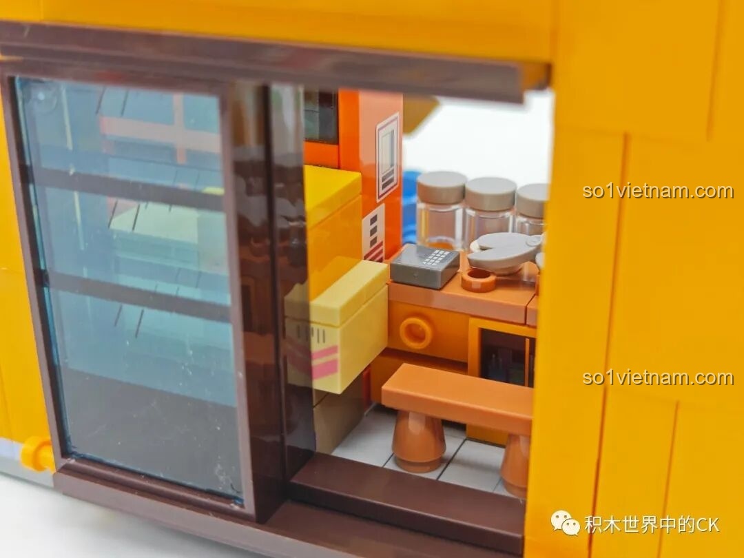 Nhìn qua ô cửa sổ trượt vào bên trong không gian của Tiệm Tạp Hóa Maruko, thấy rõ các kệ hàng và nội thất.