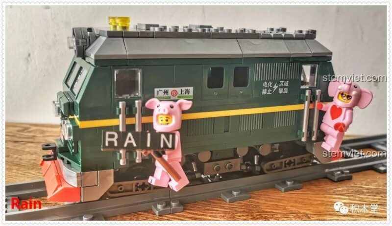 Minifigure cầm biển 'RAIN' đứng trước đầu tàu hỏa xanh lá cây Panlos Brick 610008, tạo nên khung cảnh độc đáo và hài hước.