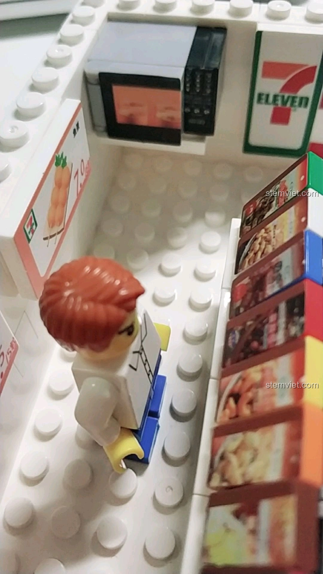 Minifigure đứng gần lò vi sóng và kệ hàng trong mô hình cửa hàng tiện lợi 7-Eleven.
