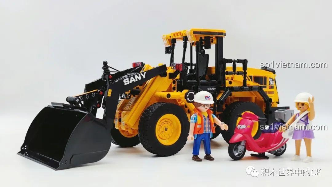 Mô hình xe xúc lật Sembo 712016 đặt cạnh các nhân vật minifigure để so sánh tỷ lệ và kích thước, thể hiện khả năng vui chơi.