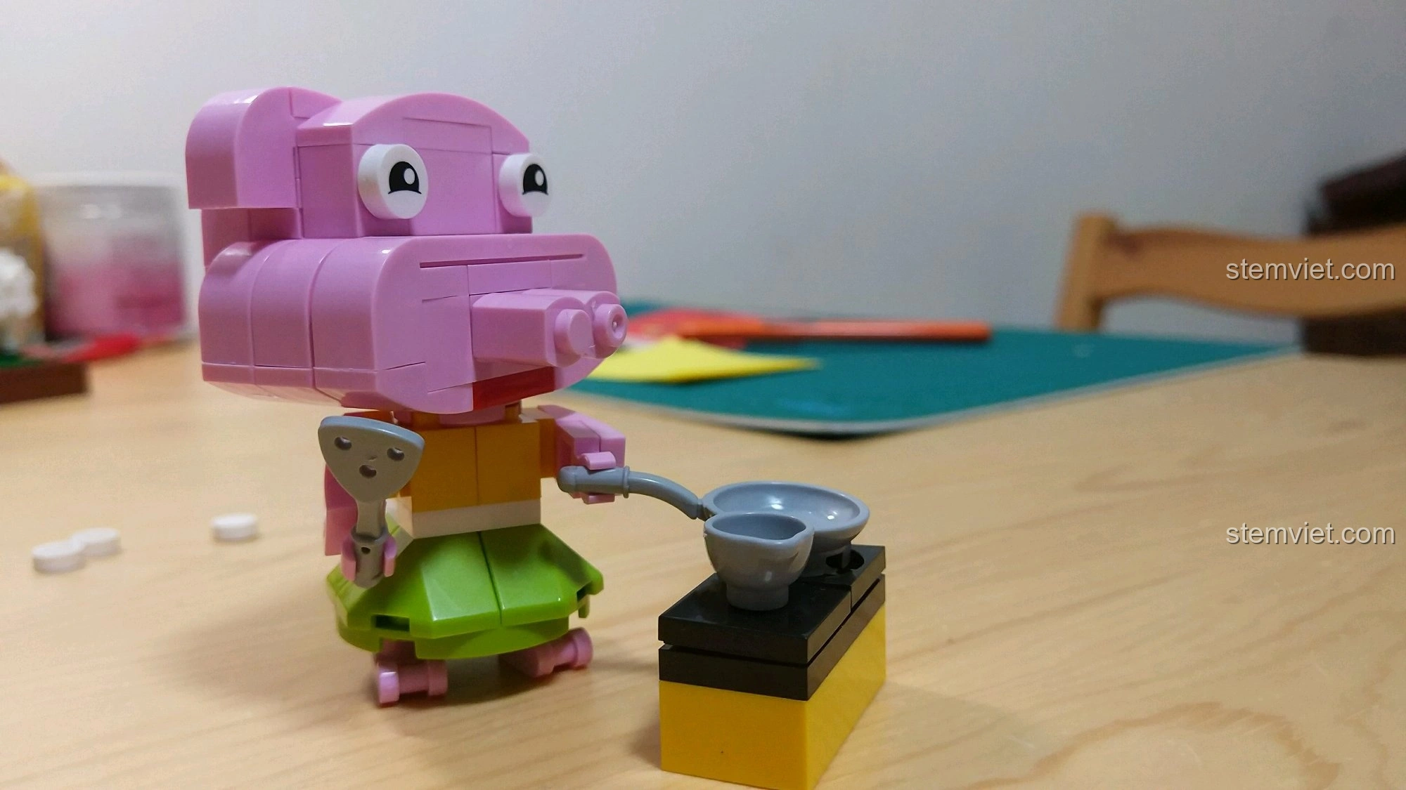 Góc nghiêng của mô hình Heo Hợi, cho thấy chi tiết váy xanh, xẻng, chảo và bếp mini, cùng biểu cảm khuôn mặt.