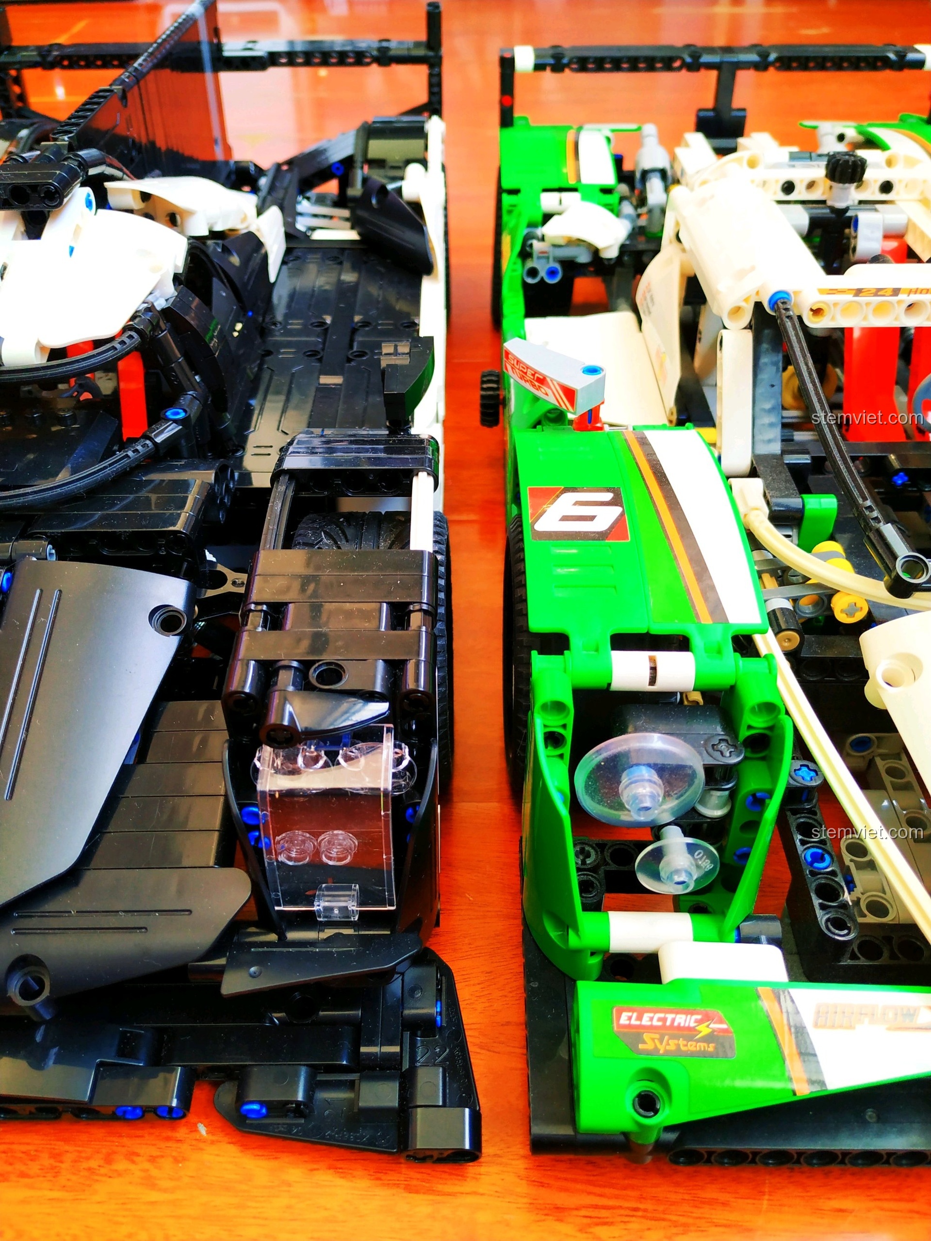 Hai phiên bản xe đua Porsche 919 Hybrid, một đen và một xanh lá, đặt cạnh nhau trên sàn gỗ.