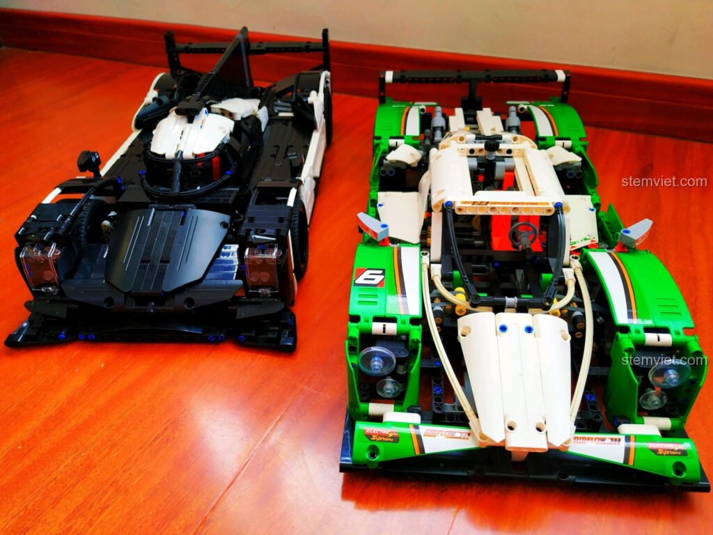 So sánh hai phiên bản xe đua Porsche 919 Hybrid, một đen và một xanh lá, đặt cạnh nhau trên sàn gỗ.