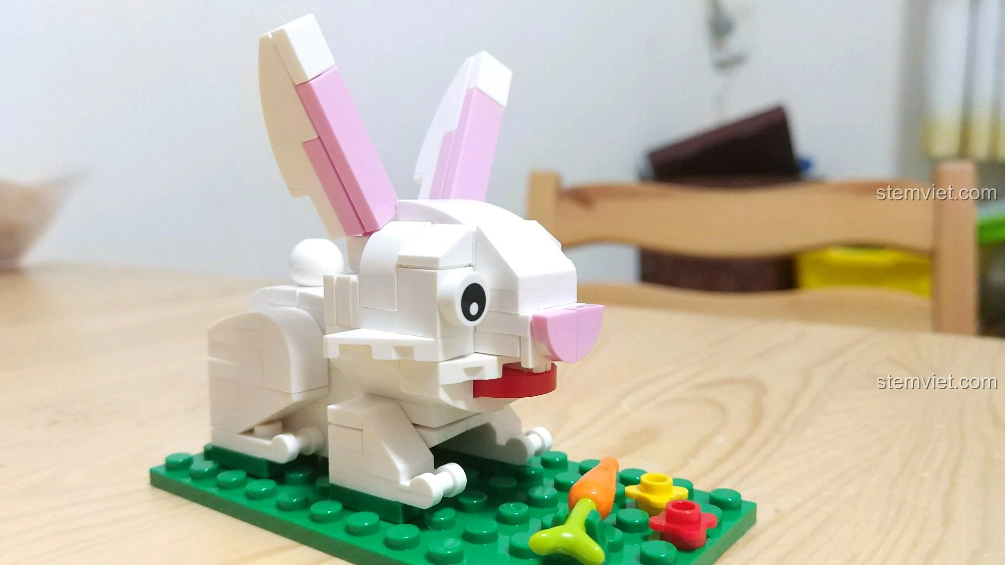 Mô hình Thỏ Mão màu trắng với tai hồng, ngồi trên nền cỏ xanh có cà rốt và hoa, từ bộ lắp ráp 12 Con Giáp XINGBAO XB-18001.