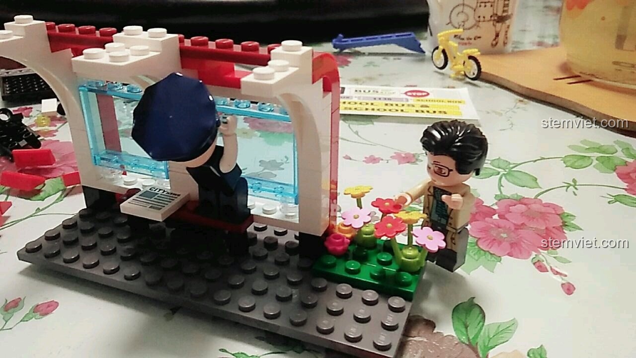 Cô giáo đang trồng hoa trang trí cho trạm chờ xe buýt đã có mái che trong bộ xếp hình QMAN 1136.