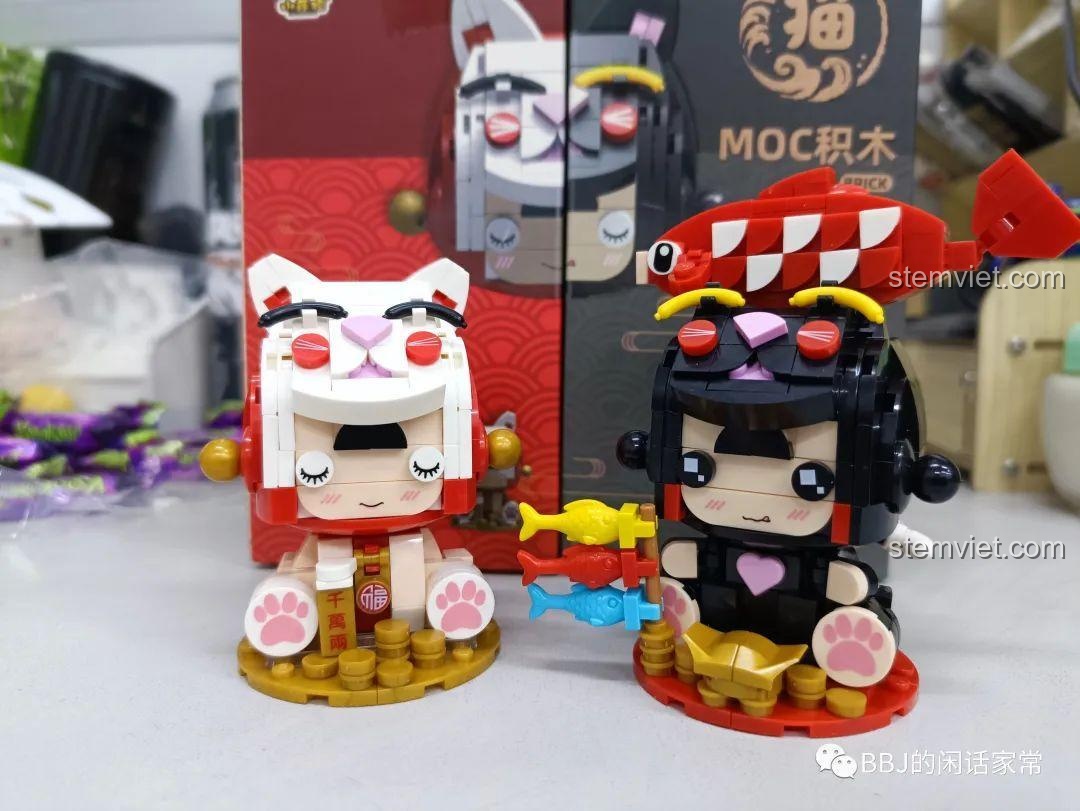 Hai mô hình Mèo Thần Tài và Cô Bé Mèo Cá Koi được trưng bày cùng nhau, phía sau là vỏ hộp sản phẩm.