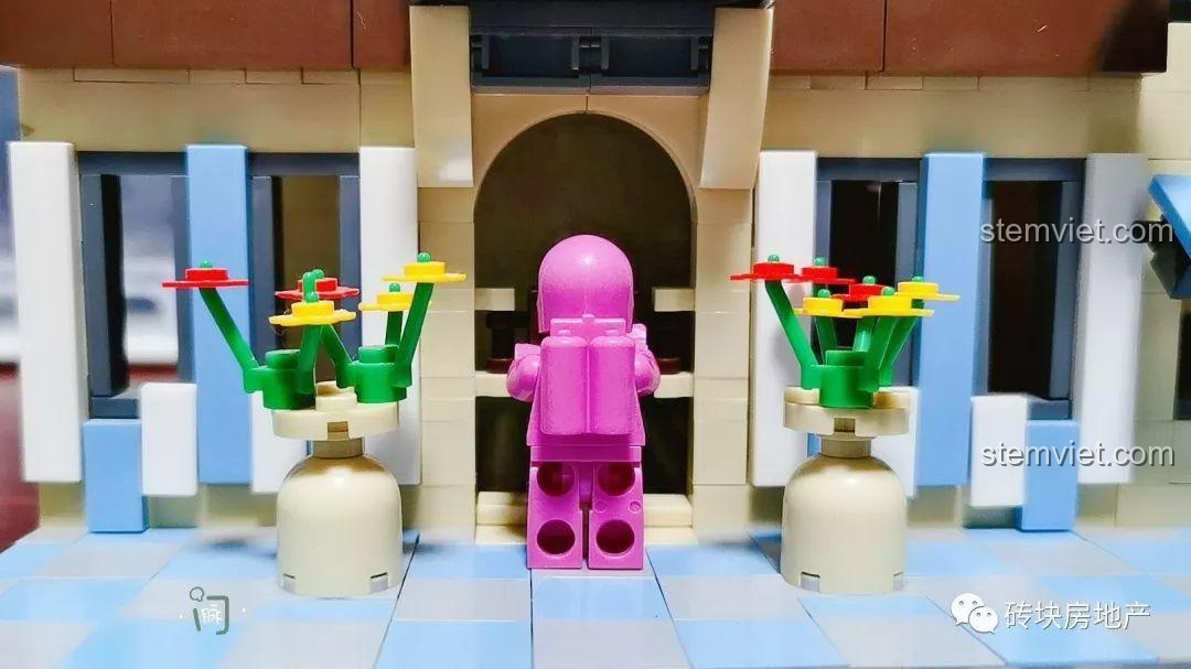 Nhân vật phi hành gia hồng đứng trước lối vào chính của Biệt Thự Đồng Quê Kiểu Pháp WANGE 5311, với hai chậu hoa trang trí.