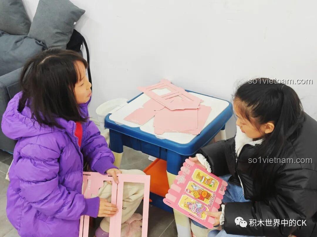 Hai bé gái đang lắp ráp kệ trưng bày bằng bìa cứng đi kèm trong bộ đồ chơi QMAN 35018.