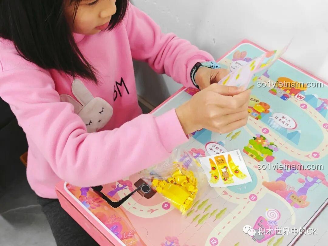 Bé gái lấy ra một túi mảnh ghép màu vàng và tờ hướng dẫn từ hộp QMAN 35018, chuẩn bị lắp ráp mô hình bánh bao.