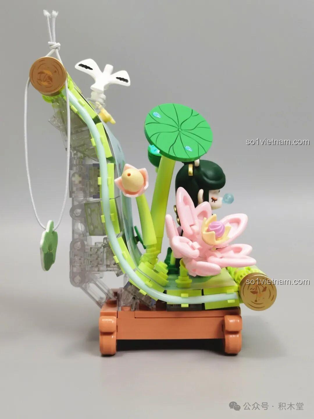 Góc nhìn chéo từ phía sau của mô hình Nanci mùa Hạ, làm nổi bật bông sen hồng và Nanci giữa khung cảnh xanh mát.