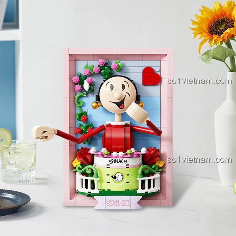 Mô hình tranh 3D Olive Oyl 86404 được trưng bày trên bàn, bên cạnh lọ hoa hướng dương, thể hiện vẻ đẹp nghệ thuật của sản phẩm.