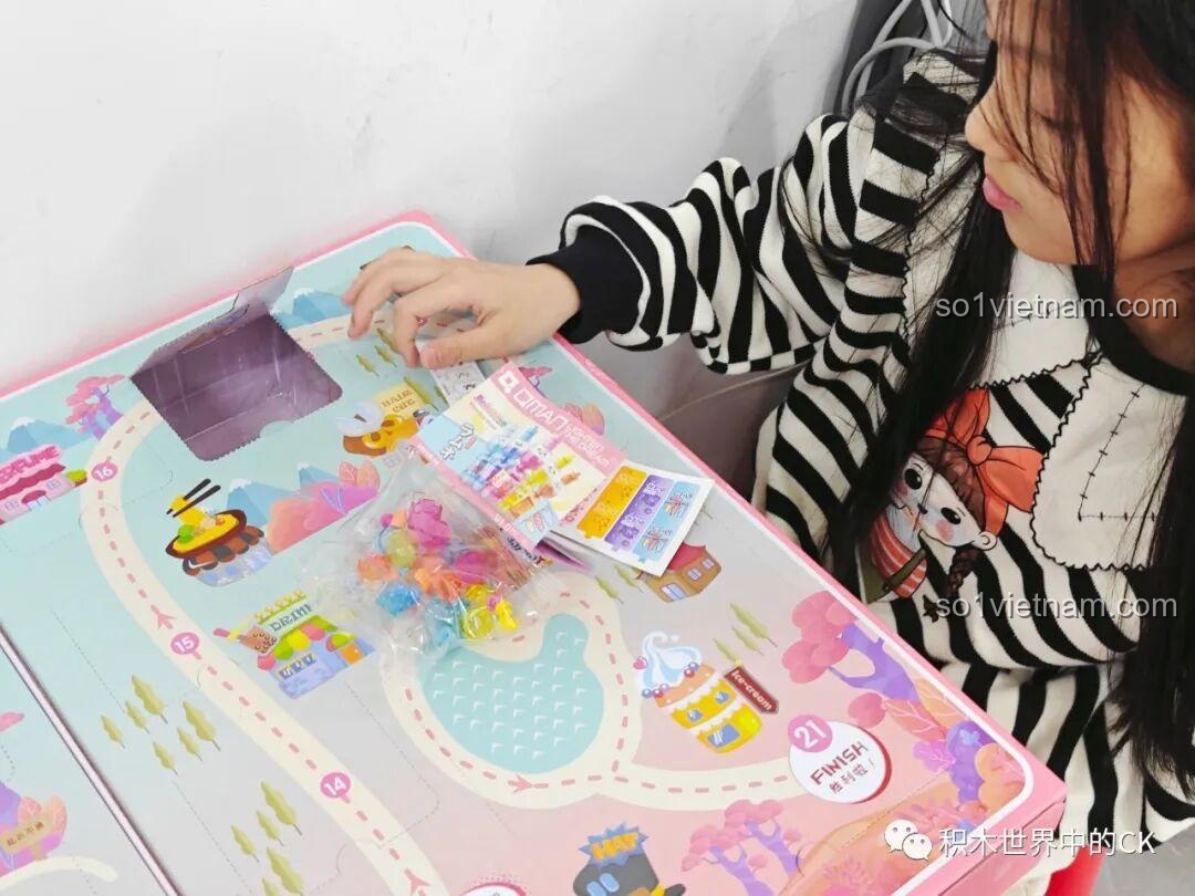 Một bé gái khác mở túi mảnh ghép nhiều màu sắc từ bộ đồ chơi QMAN 35018, háo hức khám phá mô hình tiếp theo.