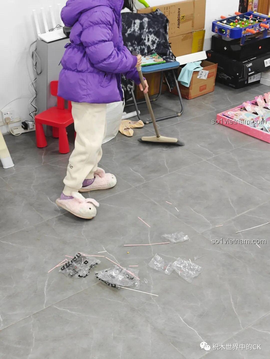 Một bé gái đang quét dọn các túi nilon sau khi chơi xong bộ lắp ráp QMAN 35018, rèn luyện tính ngăn nắp.