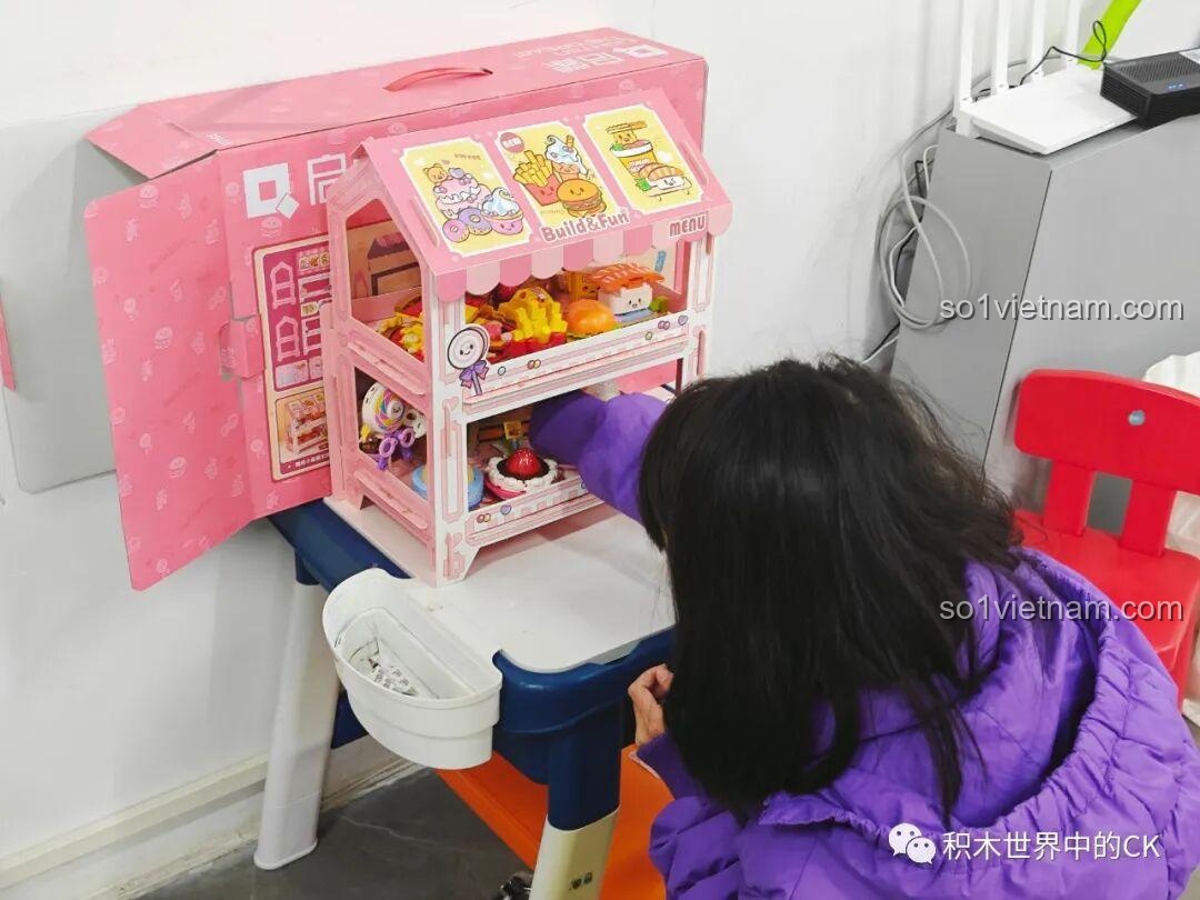 Một bé gái đang sắp xếp các mô hình món ăn mini đã lắp ráp lên kệ trưng bày của bộ QMAN 35018.